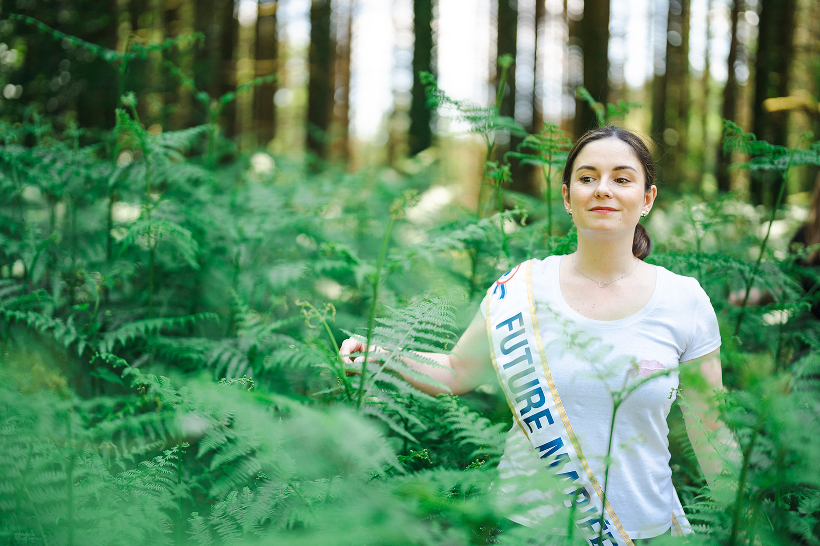 Future mariée posant au milieu des fougères lors d’une séance photo EVJF en forêt dans les Vosges, avec une écharpe “Future Mariée”.