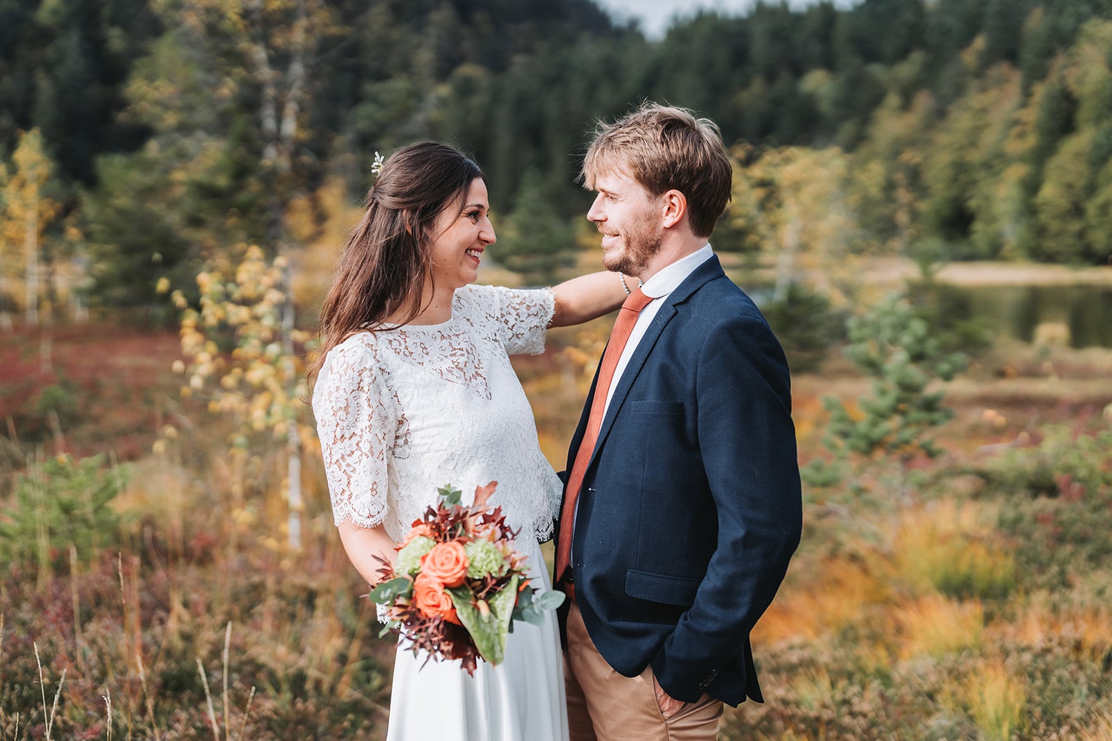 Un couple souriant lors de leur mariage en plein air, entouré de végétation automnale. La mariée porte une robe blanche en dentelle et tient un bouquet de fleurs orange et vertes, tandis que le marié porte un costume bleu marine avec une cravate orange.
