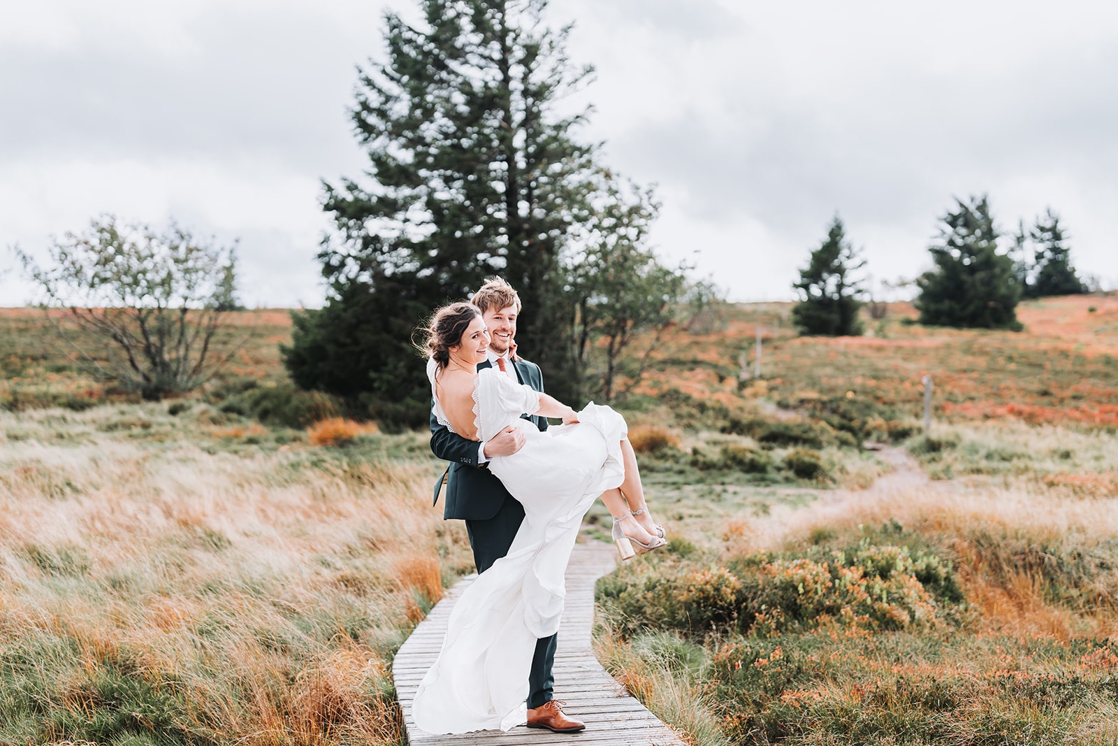 Un marié porte sa mariée dans ses bras sur un chemin en bois au milieu d’un paysage naturel. Le couple sourit, entouré de prairies et d’arbres sous un ciel nuageux.
