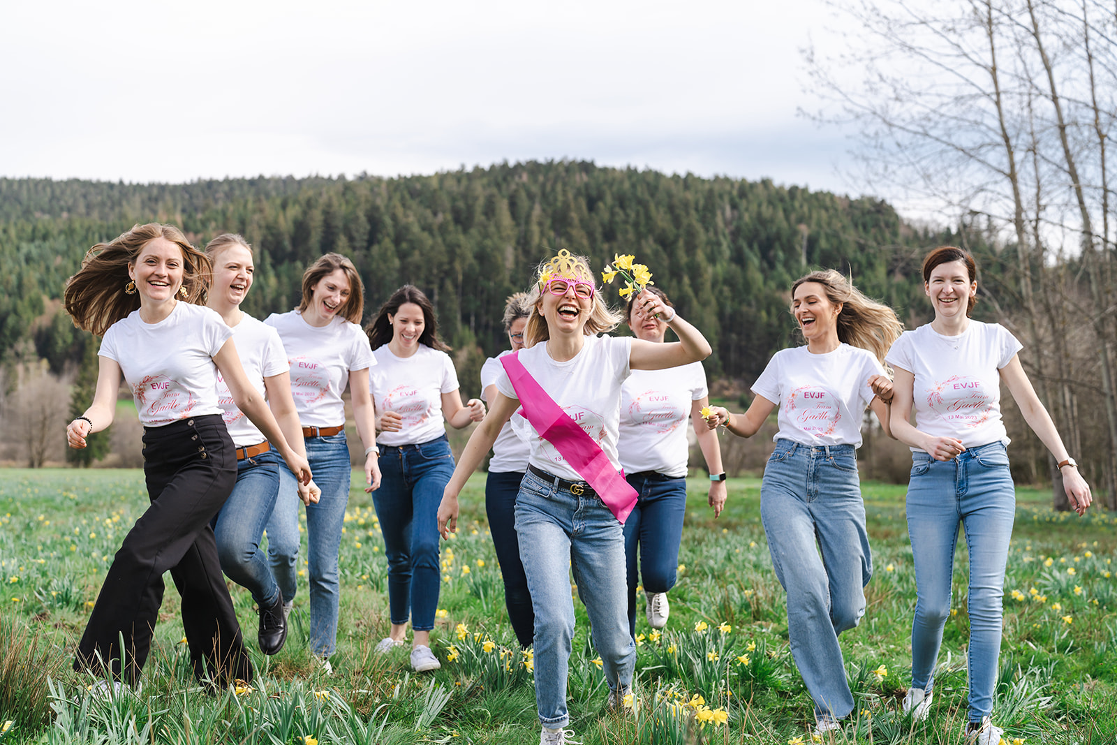 Groupe de femmes riant et courant dans un champ fleuri lors d’une séance photo EVJF dans les Vosges, avec la future mariée en avant-plan portant une couronne, une écharpe rose et des fleurs.